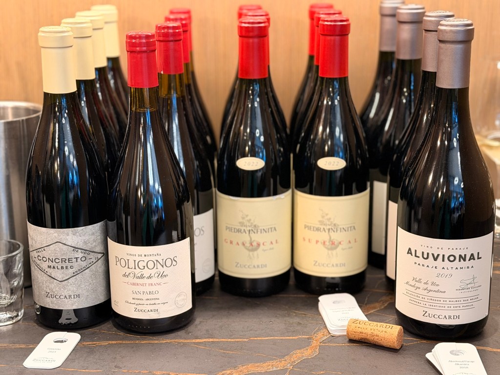 A display of various bottles of Zuccardi red wines, including Concreto Malbec, Polígonos Cabernet Franc, and Aluvional, arranged on a table with wine corks and tags.