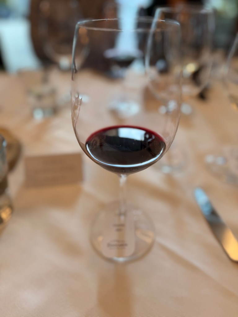 A glass of red wine on a dining table, surrounded by other glassware and utensils.