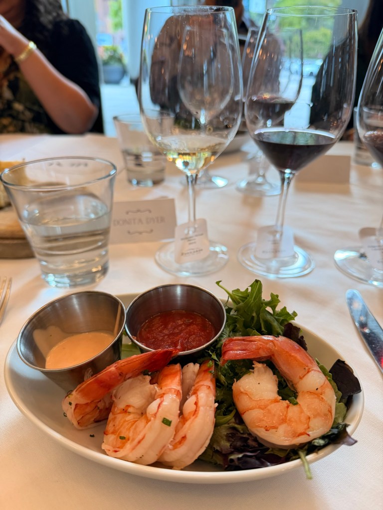 A plate of shrimp served on a bed of mixed greens, accompanied by two dipping sauces in small metal bowls, one beige and one red. Several wine glasses are visible in the background.