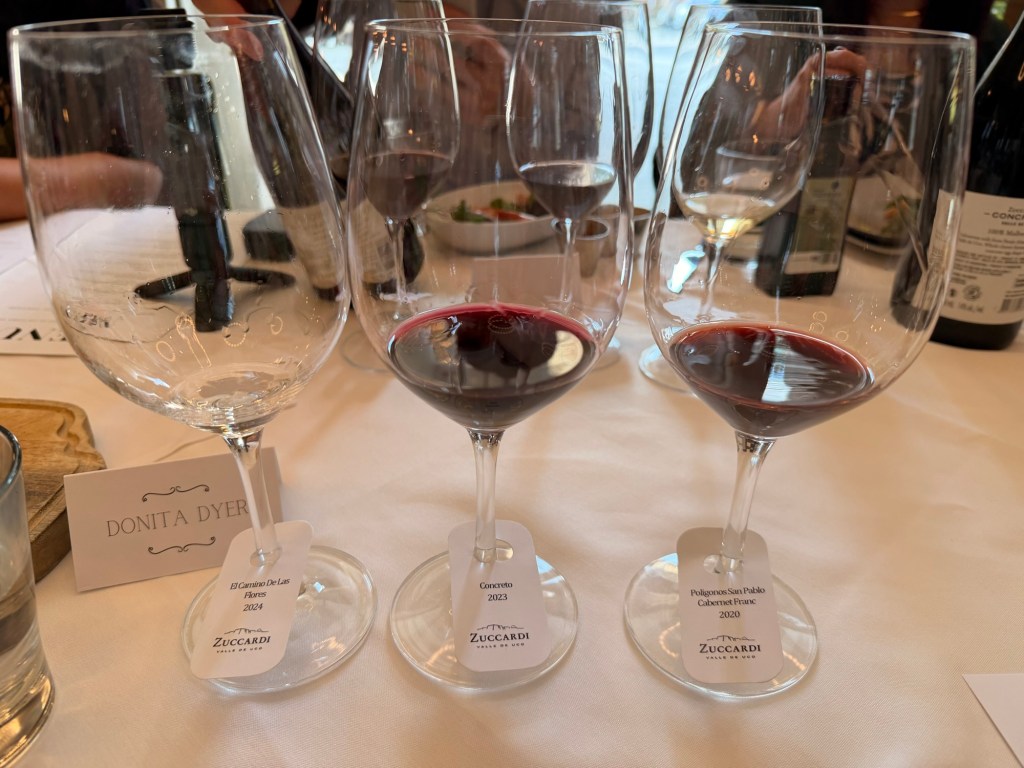 Three wine glasses filled with red wine, each labeled with wine names and vintages, placed on a white tablecloth. A name card labeled 'DONITA DYER' is also visible.