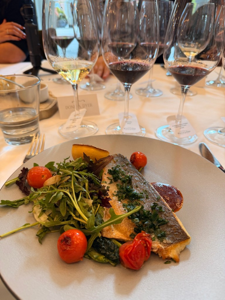A plated dish featuring a piece of fish topped with herbs, accompanied by roasted tomatoes, a slice of lemon, and a bed of mixed greens, with several wine glasses in the background.