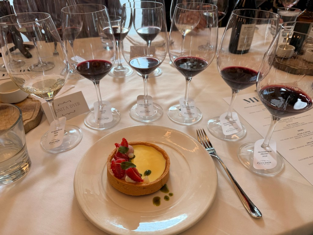 A dining table set with several wine glasses, including red and white wines, alongside a dessert plate featuring a tart topped with strawberries and cream, garnished with green sauce.