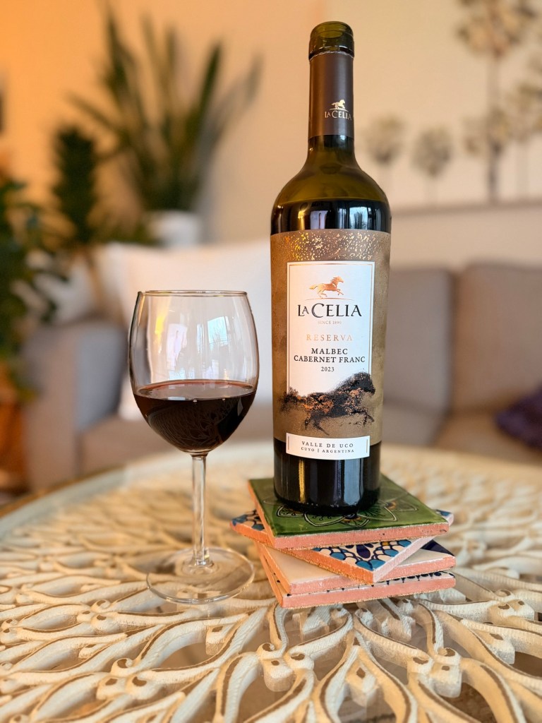 A bottle of La Celia Reserva Malbec Cabernet Franc wine next to a filled wine glass, placed on a decorative table with stacked coasters. The background features soft furnishings and plants, creating a cozy atmosphere.
